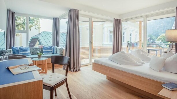 A modern hotel room with a large bed, a desk, and a seating area with a view of the outside. - Hotel Kendler (St. Gilgen)
