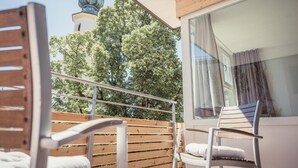 A balcony with wooden railings, a white cushioned chair, and a view of a building with a dome and trees in the background. - Hotel Kendler (St. Gilgen)