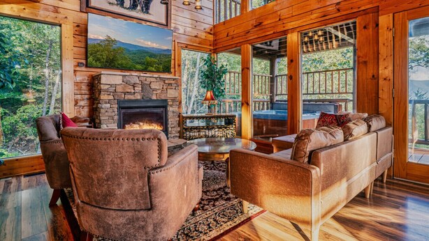 A cozy cabin interior with a stone fireplace, leather sofas, and large windows offering views of the forest. - Perfect Smokies Escape - Park View, Hot Tub & More (Sevierville)