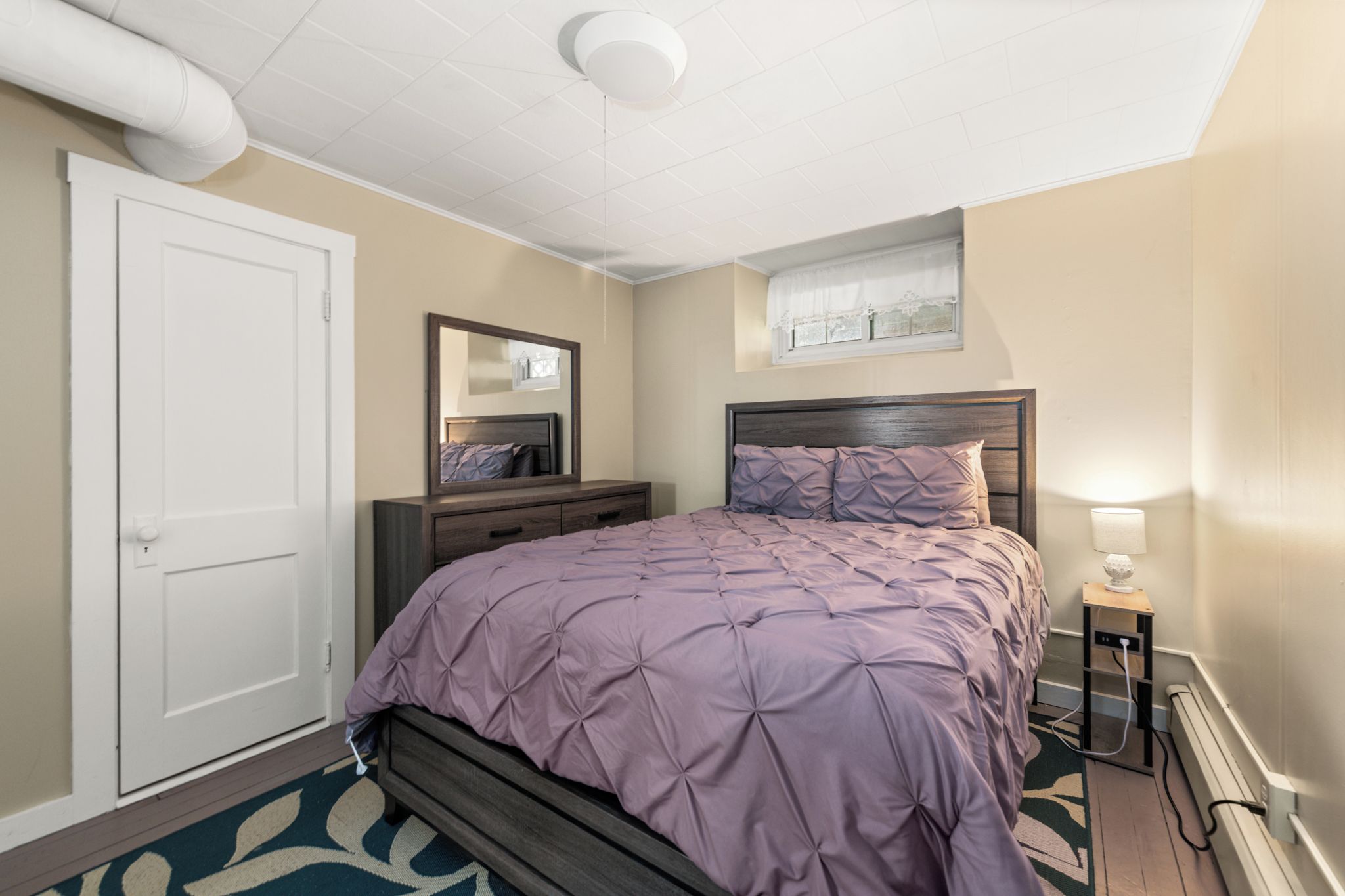 A bedroom with a bed, dresser, mirror, and a small table with a lamp.