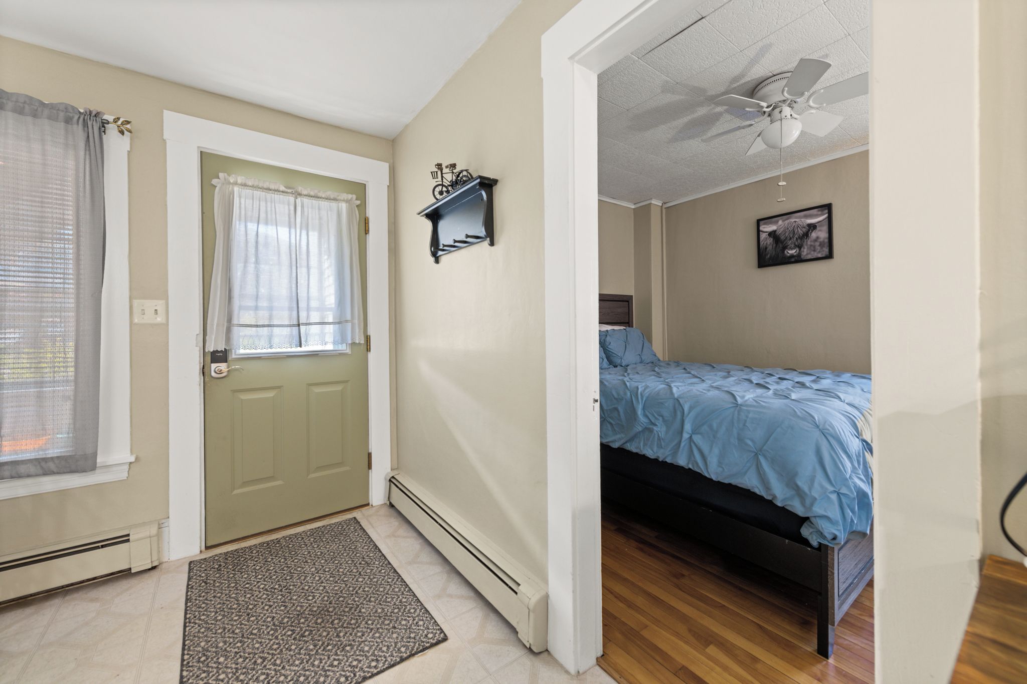 A bedroom with a bed, a door, a window with curtains, a radiator, a ceiling fan, and a wall-mounted picture.