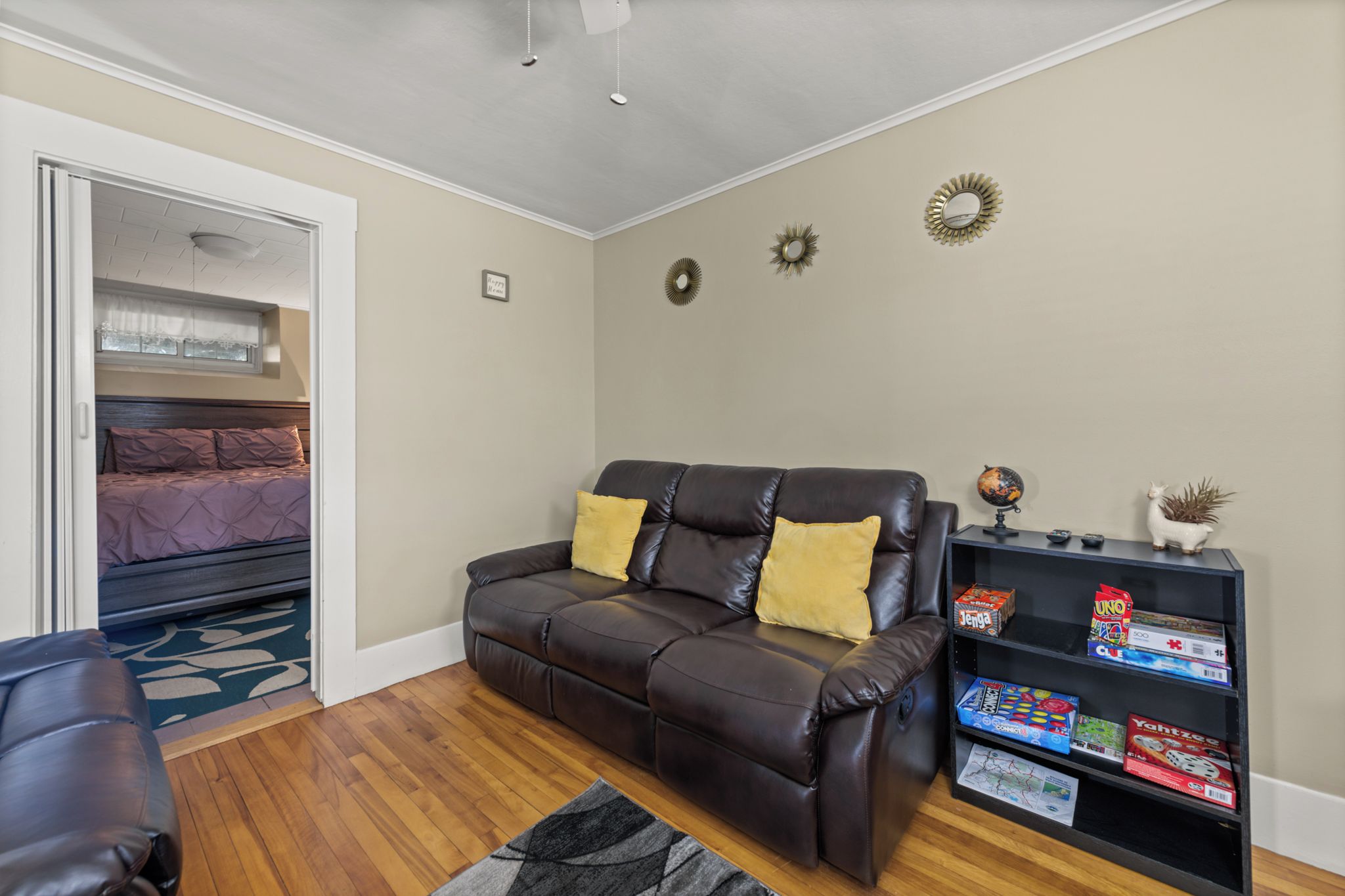 A living room with a leather sofa, a bookshelf with board games, and a bed in the adjacent room.