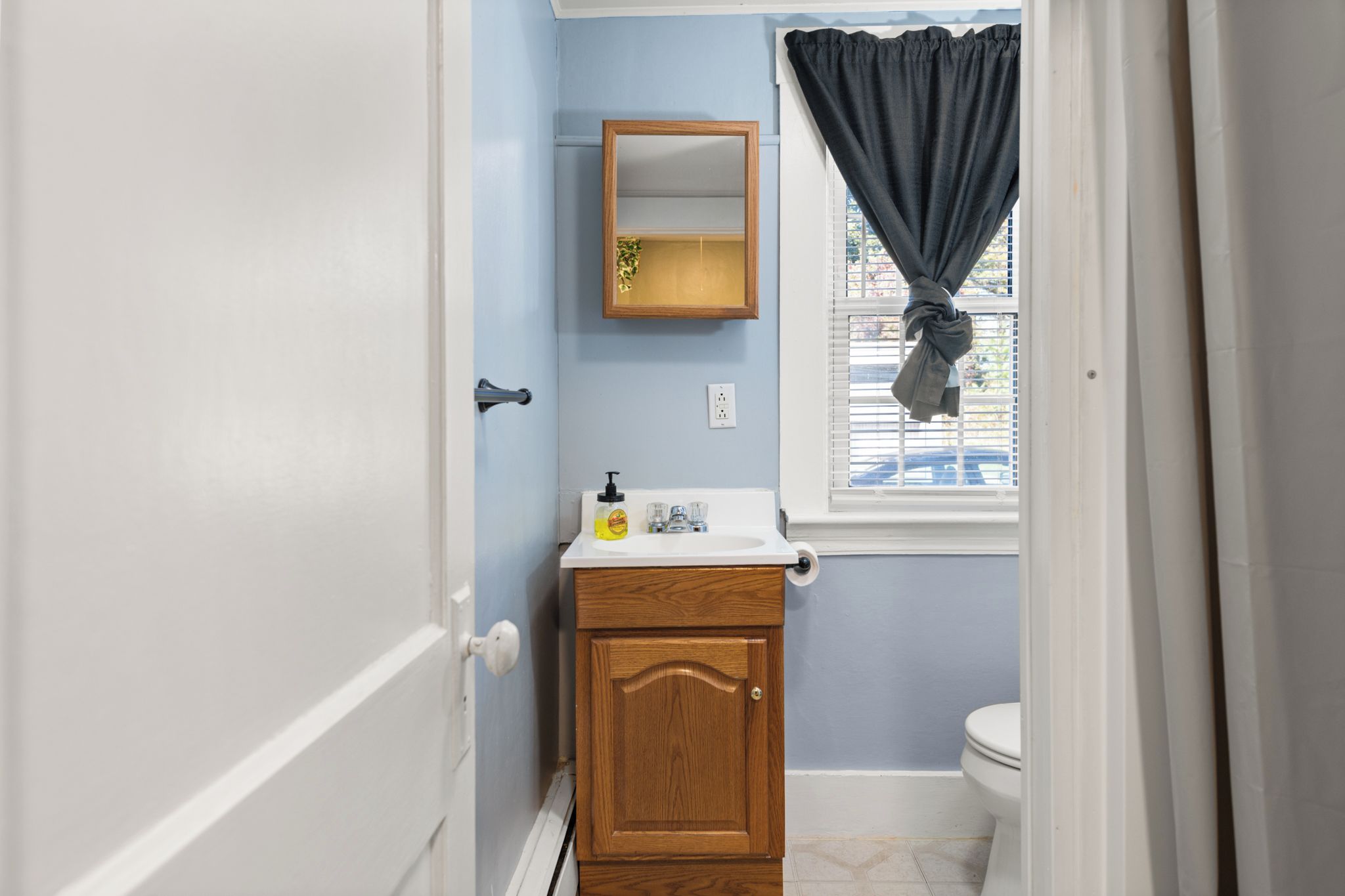 A small bathroom with a wooden cabinet, a mirror, a window with a curtain, and a toilet.