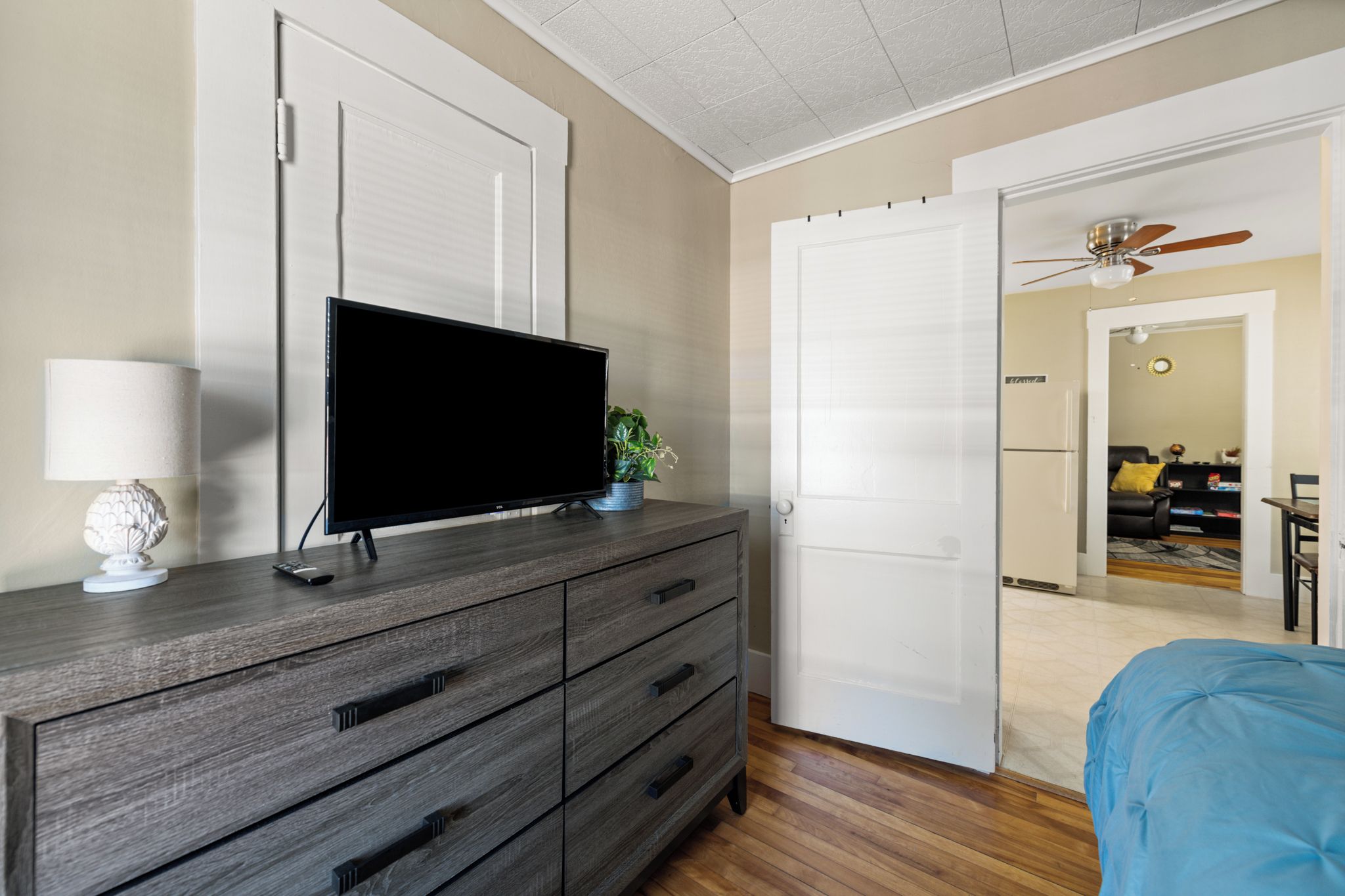 A bedroom with a TV on a dresser, a bed with a blue cover, and a door leading to a living area.