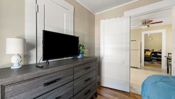 A bedroom with a TV on a dresser, a bed with a blue cover, and a door leading to a living area.