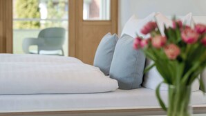 Una cama con almohadas y un jarrón de flores en una habitación con ventana y una silla al fondo.