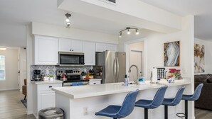 Een moderne keuken met witte kastjes, een roestvrijstalen koelkast, een magnetron en een koffiezetapparaat. Er staan drie blauwe barstoelen bij het eiland.