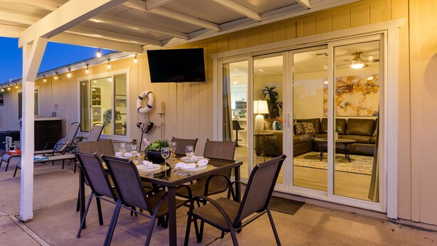 Een terras buiten met een eettafel voor vier personen, omringd door stoelen en een televisie die aan de muur is bevestigd.