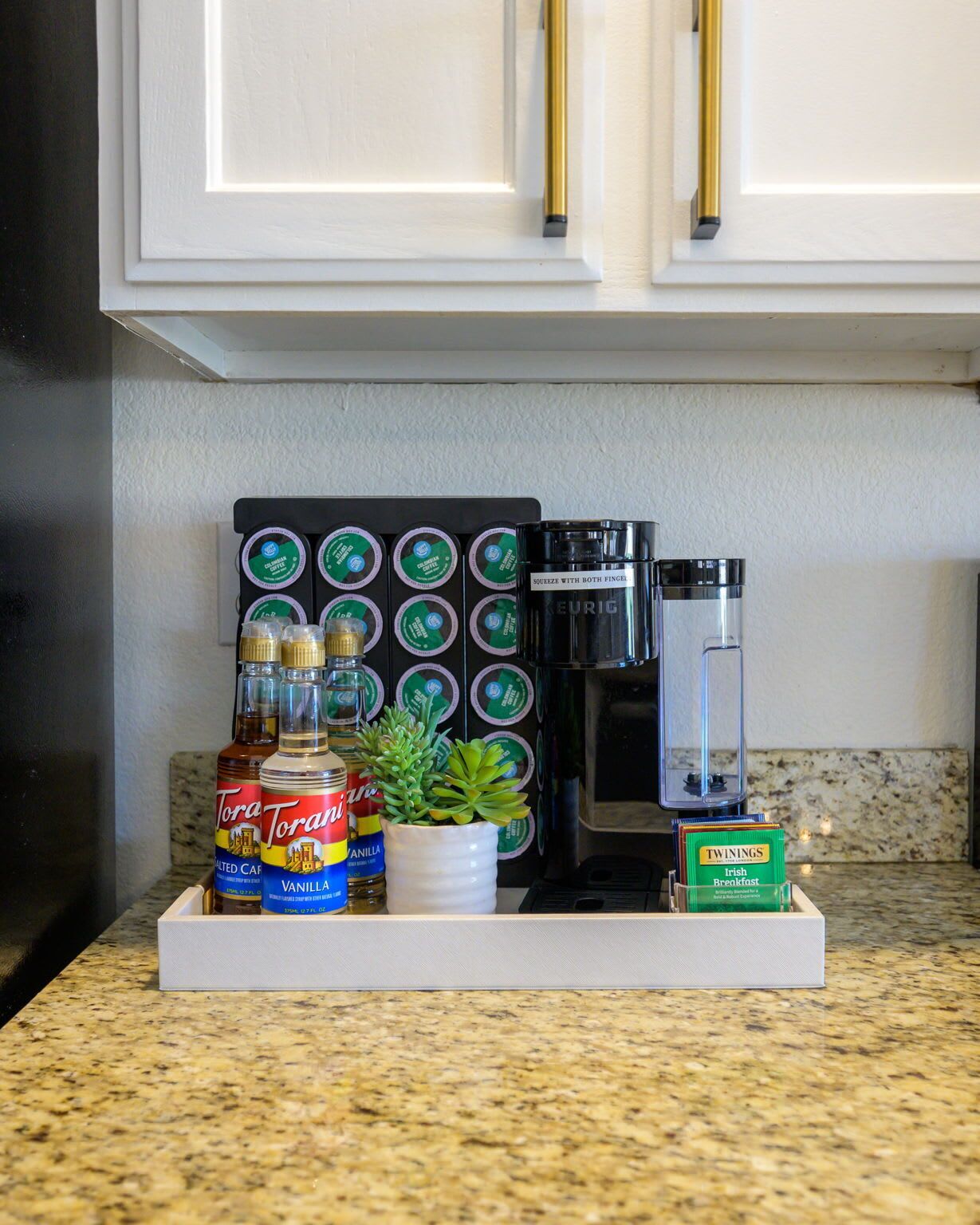 Eine Küchenarbeitsplatte mit Kaffeemaschine, einem Keurig-System, Flaschen mit Torani-Sirup, einer Topfpflanze und einer Packung Twinings Irish Breakfast Tee.