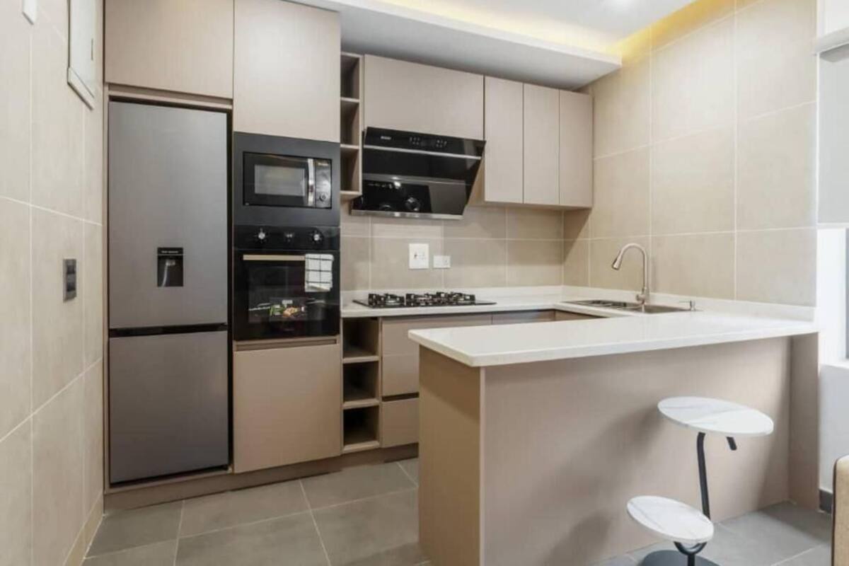 A modern kitchen with stainless steel appliances, a built-in oven, microwave, and gas stove. There are two white bar stools and a small island with a sink.
