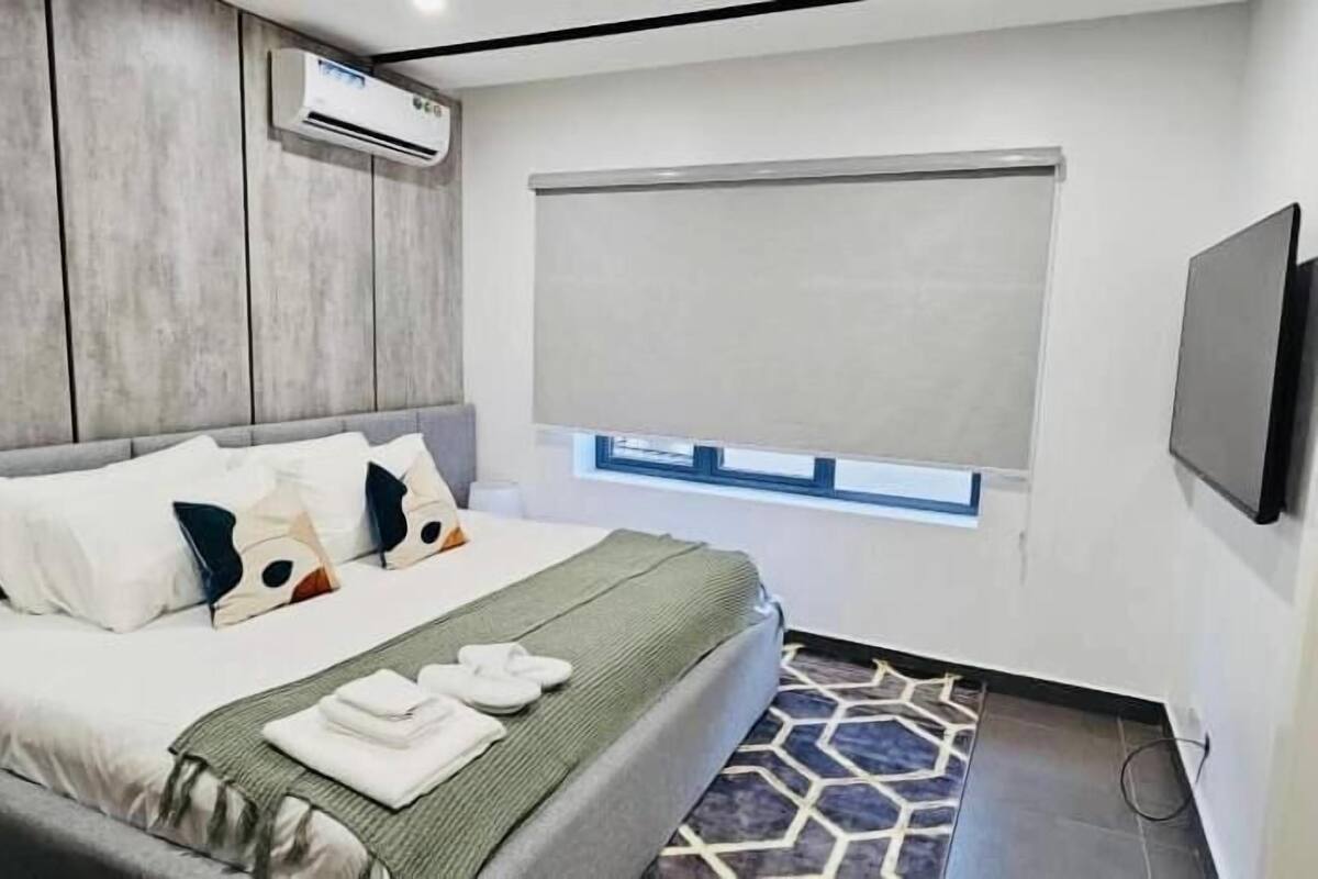 A modern bedroom with a bed, pillows, a blanket, a window, and a wall-mounted air conditioner.
