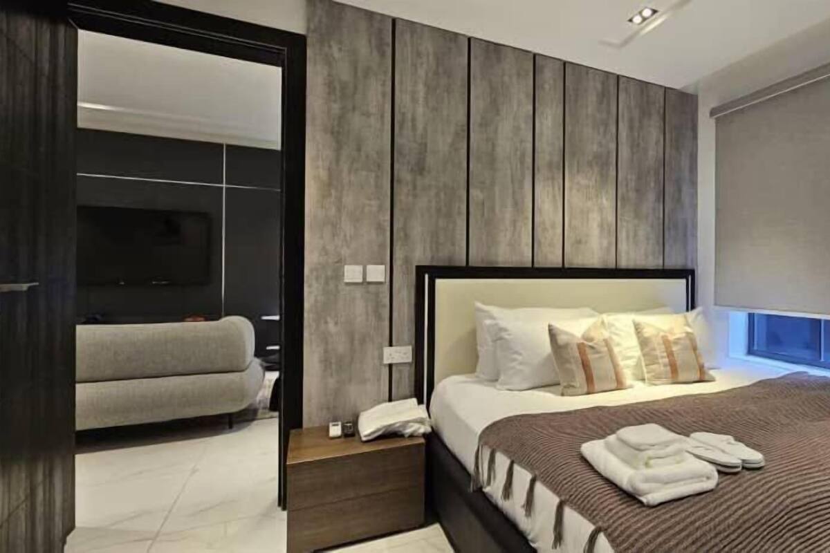 A modern bedroom with a large bed, a nightstand, a sofa, and a window with blinds.