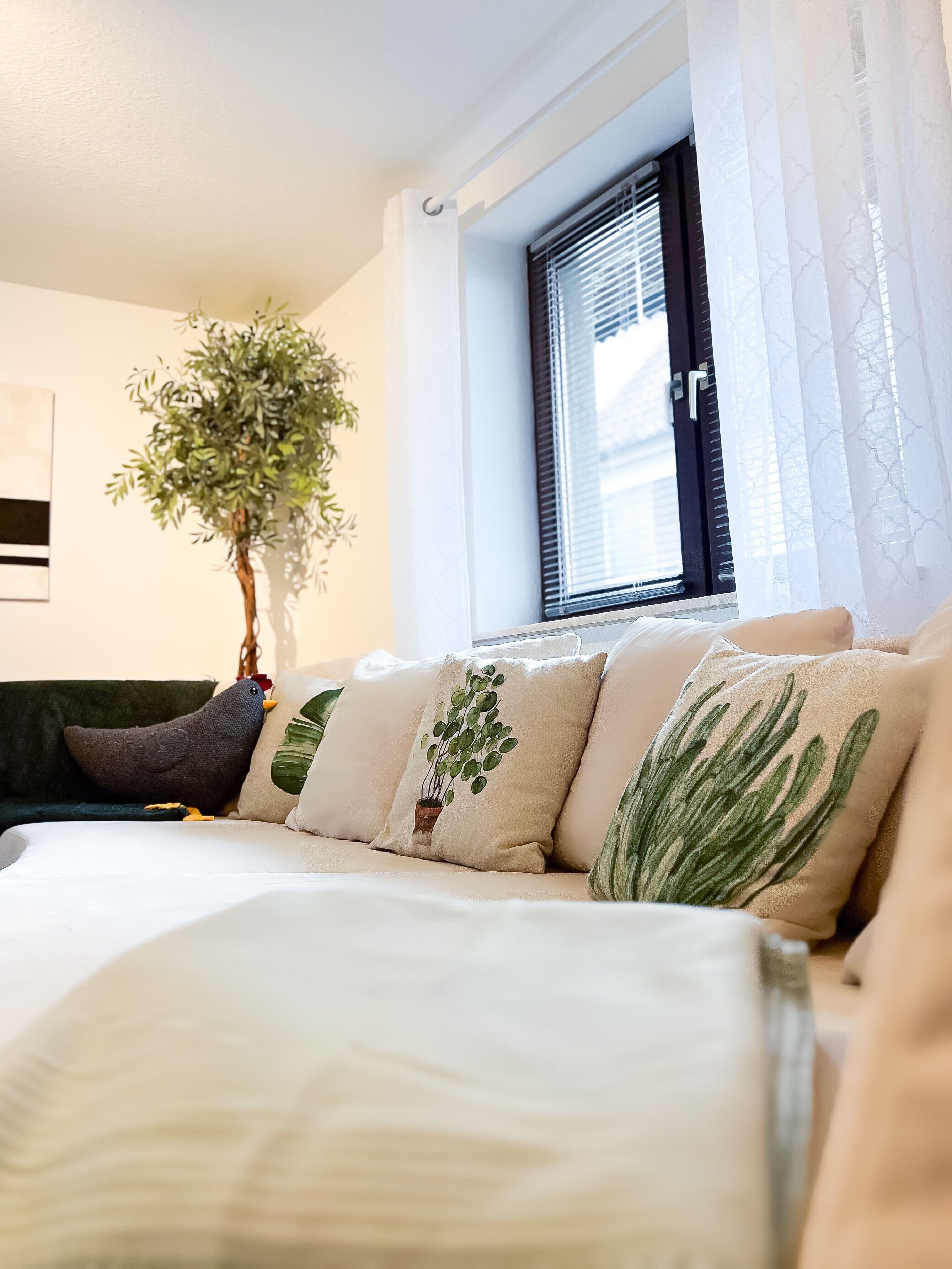 Ein Schlafzimmer mit Bett, einem Fenster mit Jalousien, einer Zimmerpflanze und einem an der Wand hängenden Kunstwerk.