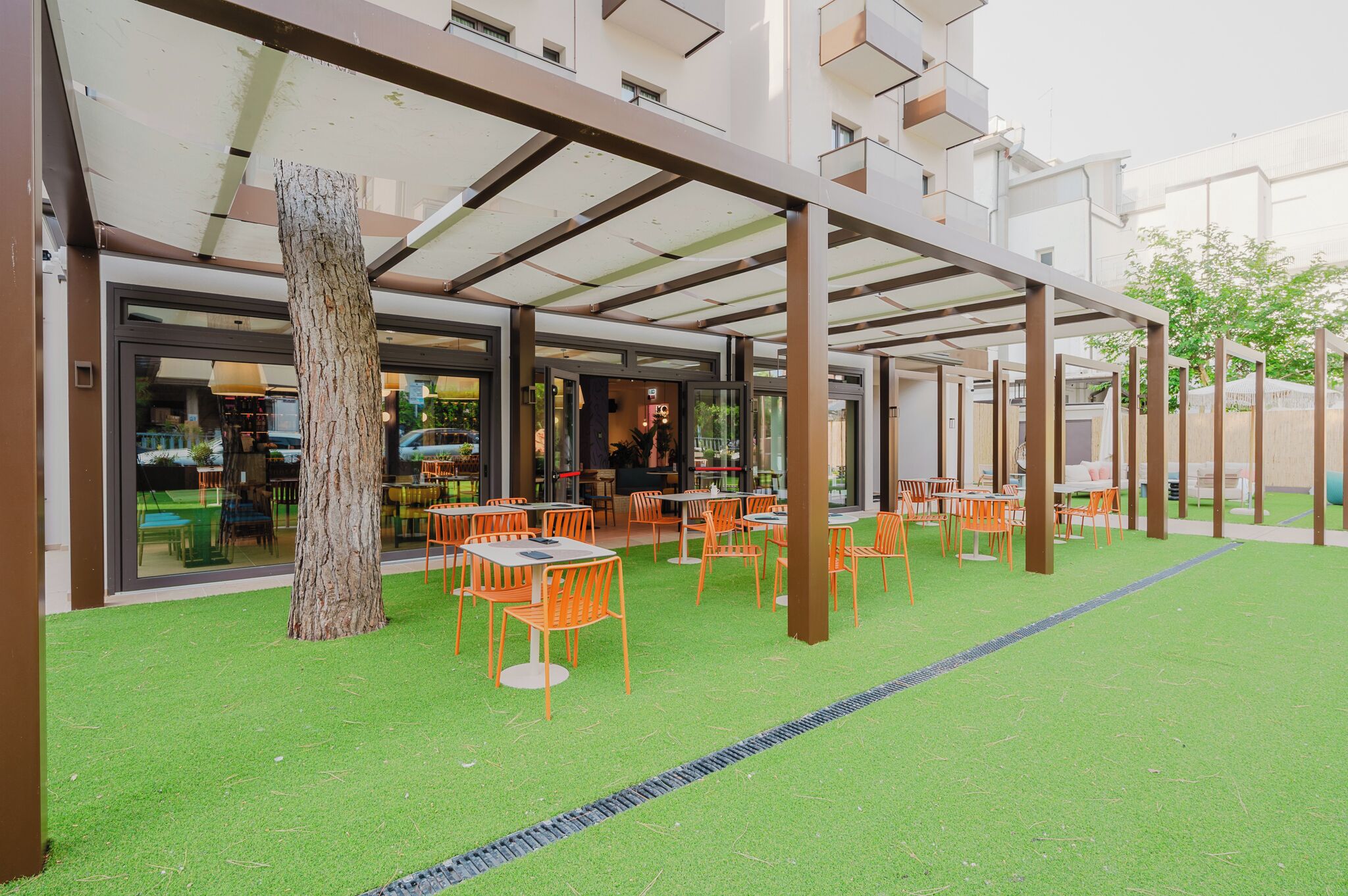 An outdoor seating area with tables and chairs, a tree, and a building entrance.