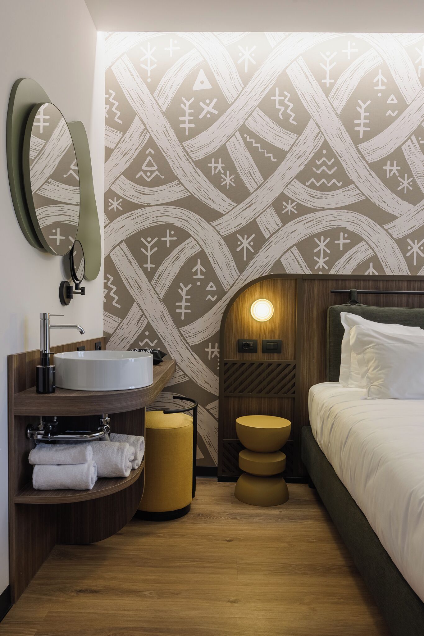 Una habitación de hotel moderna con un espejo redondo, un lavabo blanco, una cómoda de madera, una cama con ropa de cama blanca y una pared con estampado.