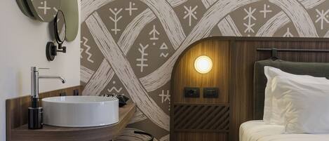 Una habitación de hotel moderna con un espejo redondo, un lavabo blanco, una cómoda de madera, una cama con ropa de cama blanca y una pared con estampado.