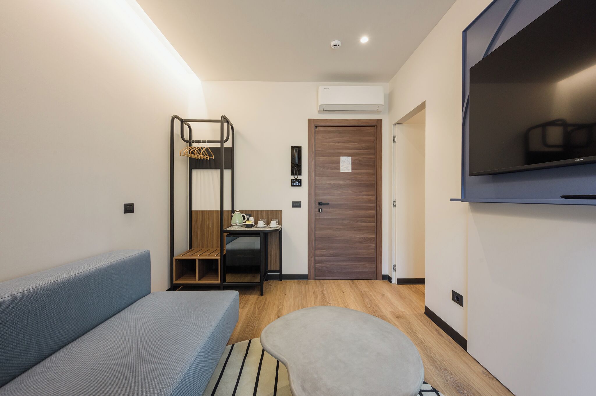 A small, modern room with a sofa, a small table with a kettle, a TV, and a wooden door.