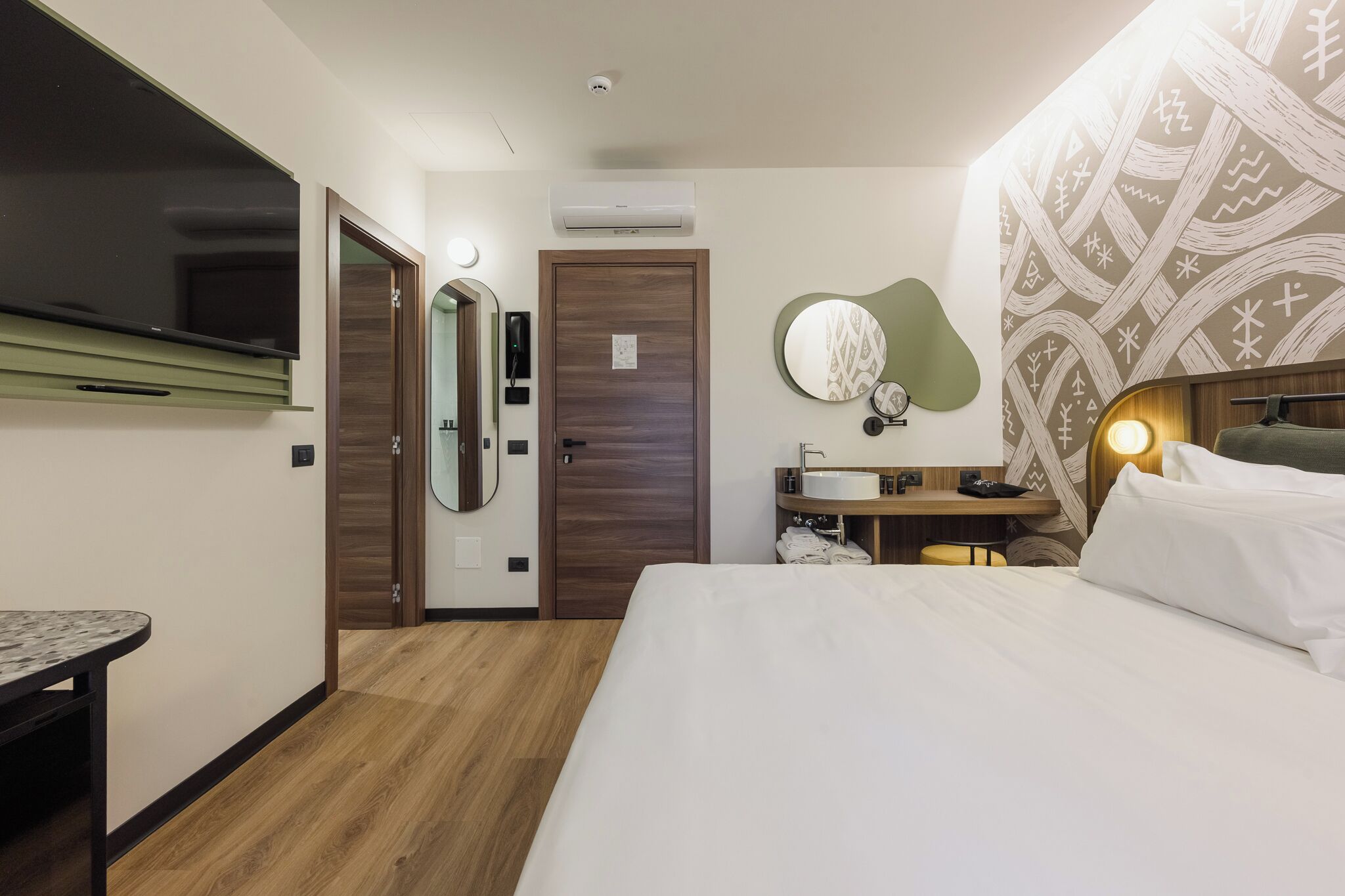 A hotel room with a bed, a desk, a mirror, a wall-mounted TV, a bathroom with a sink, and a patterned wall.