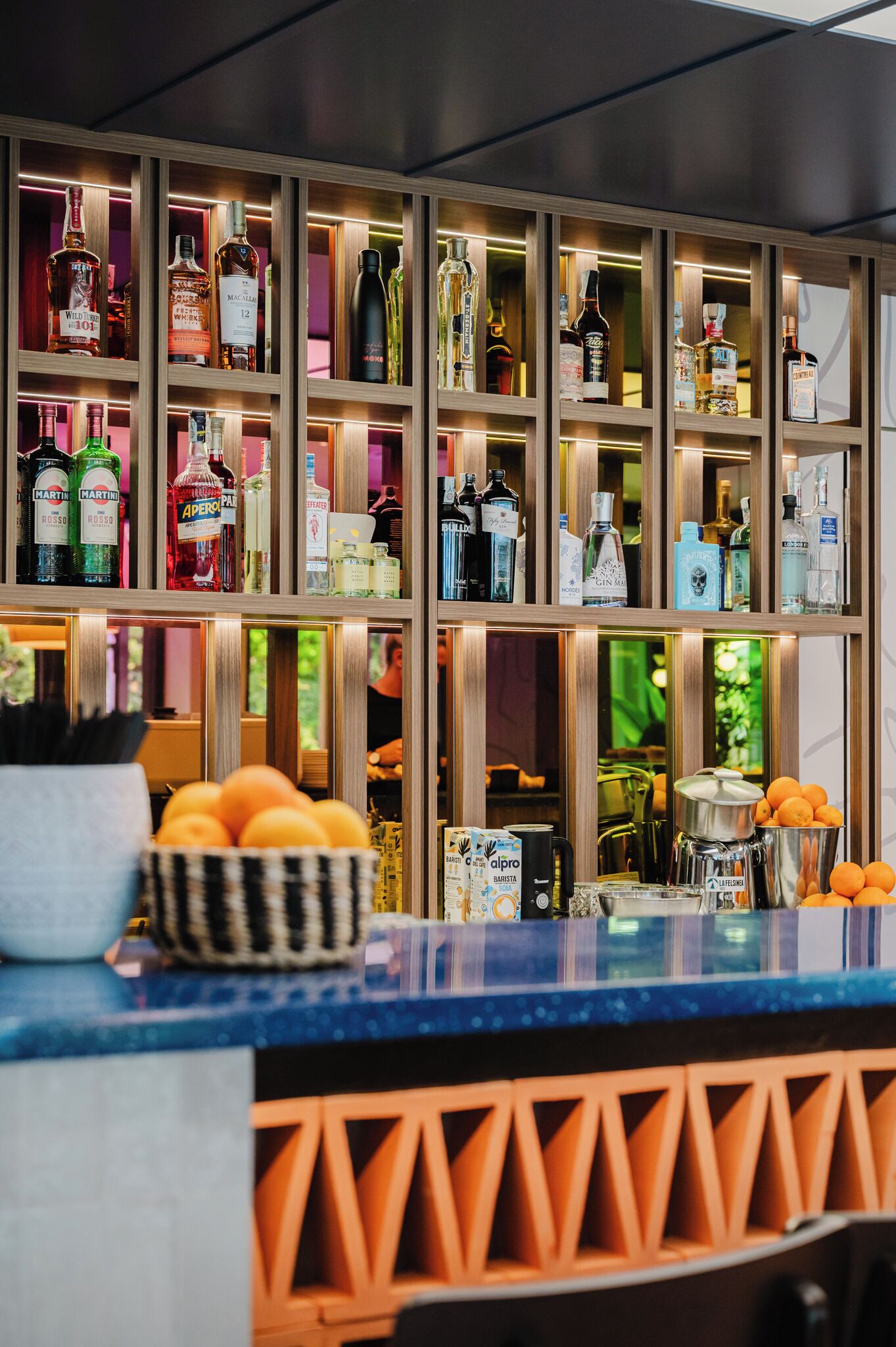 A bar area with multiple shelves of liquor bottles, a fruit bowl, and bar tools.
