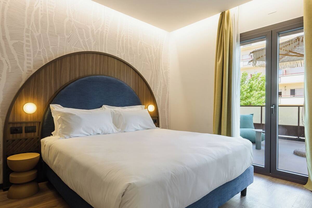 Une chambre d’hôtel moderne avec un grand lit, un plancher en bois et une vue sur l’extérieur, visible à travers une porte vitrée.