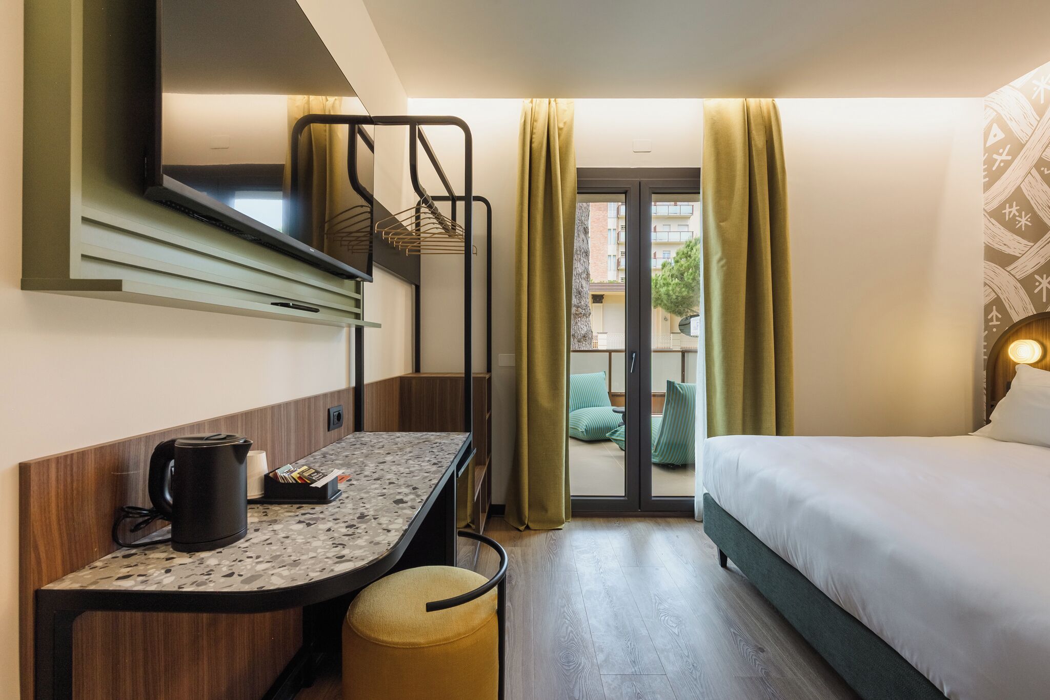 A compact hotel room with a bed, a desk with a kettle, a small stool, a TV, and a balcony with curtains.