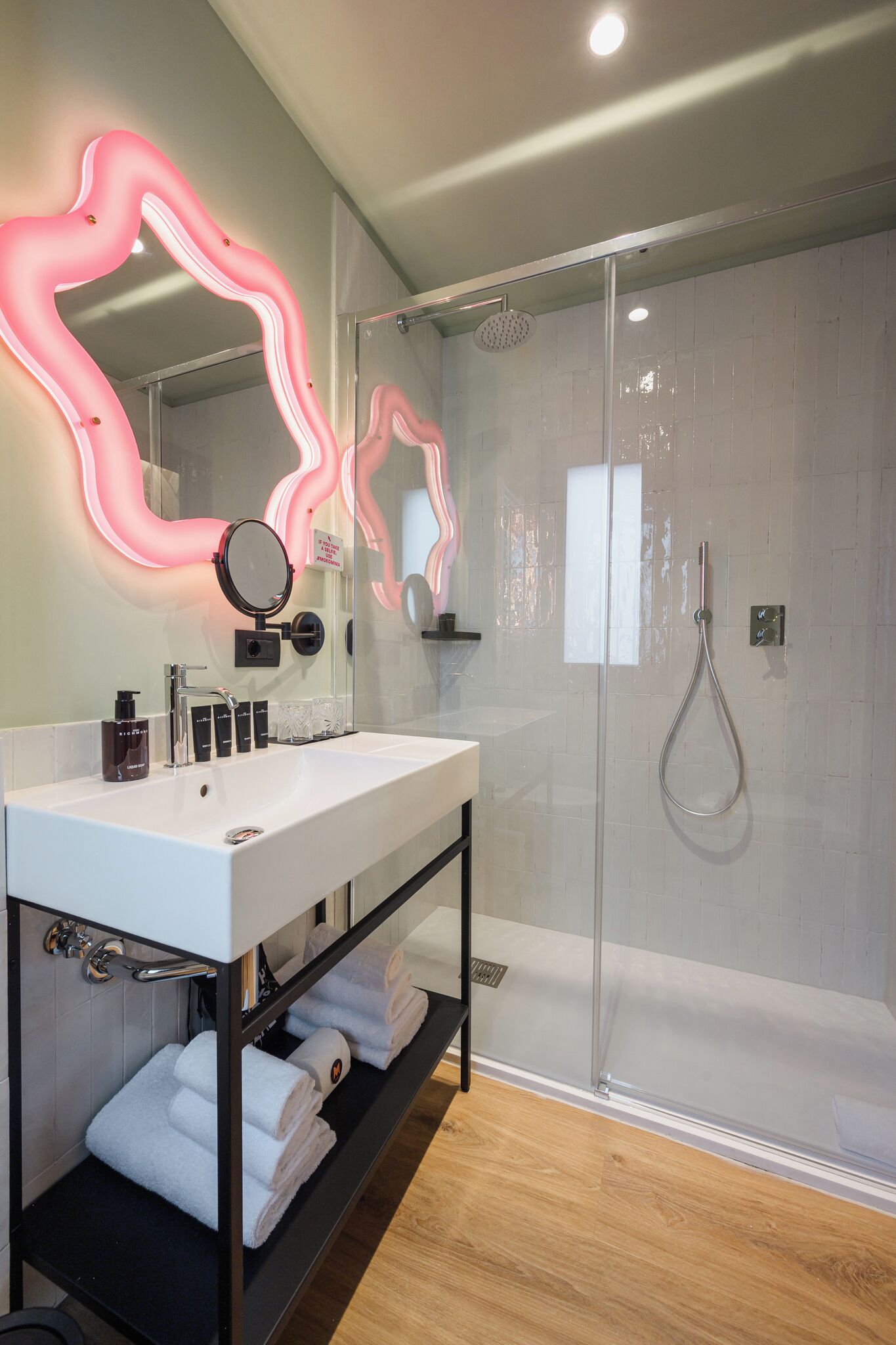 A modern bathroom with a glass shower enclosure, a white sink with a mirror, and a shelf with towels.