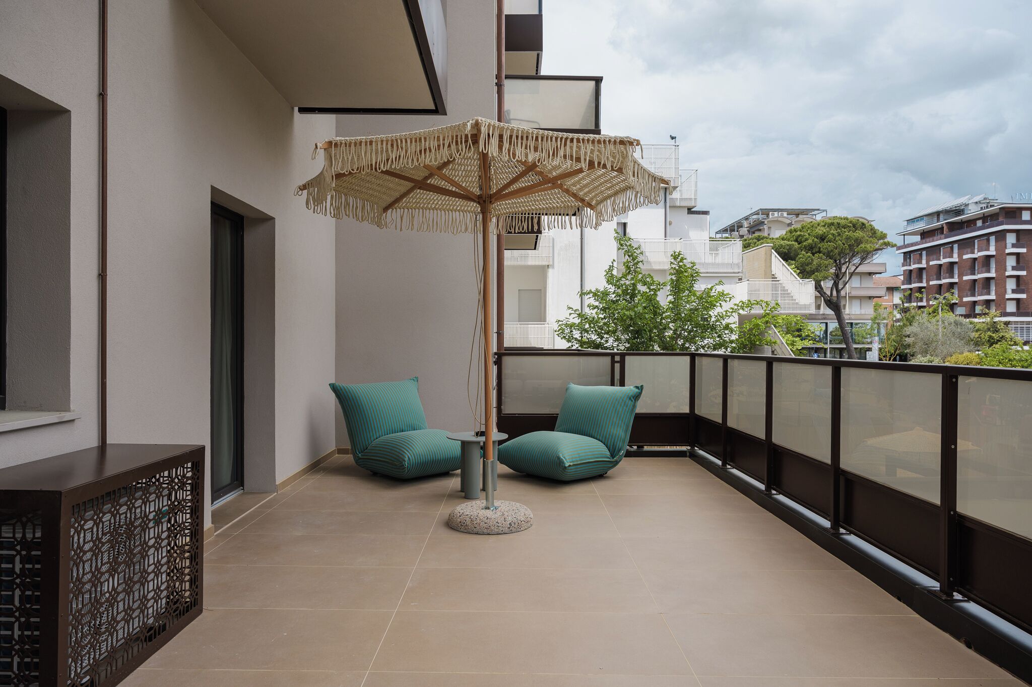 A balcony with a woven umbrella, two teal cushioned chairs, a small round table, and a decorative screen.