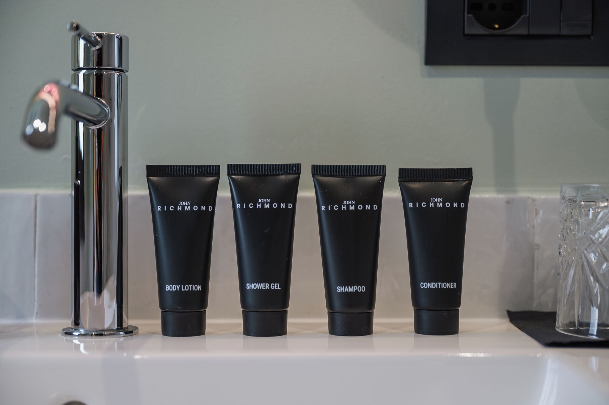 A bathroom with a chrome faucet, four black tubes labeled 'JOHN RICHMOND BODY LOTION,' 'SHOWER GEL,' 'SHAMPOO,' and 'CONDITIONER,' and a clear glass on the counter.