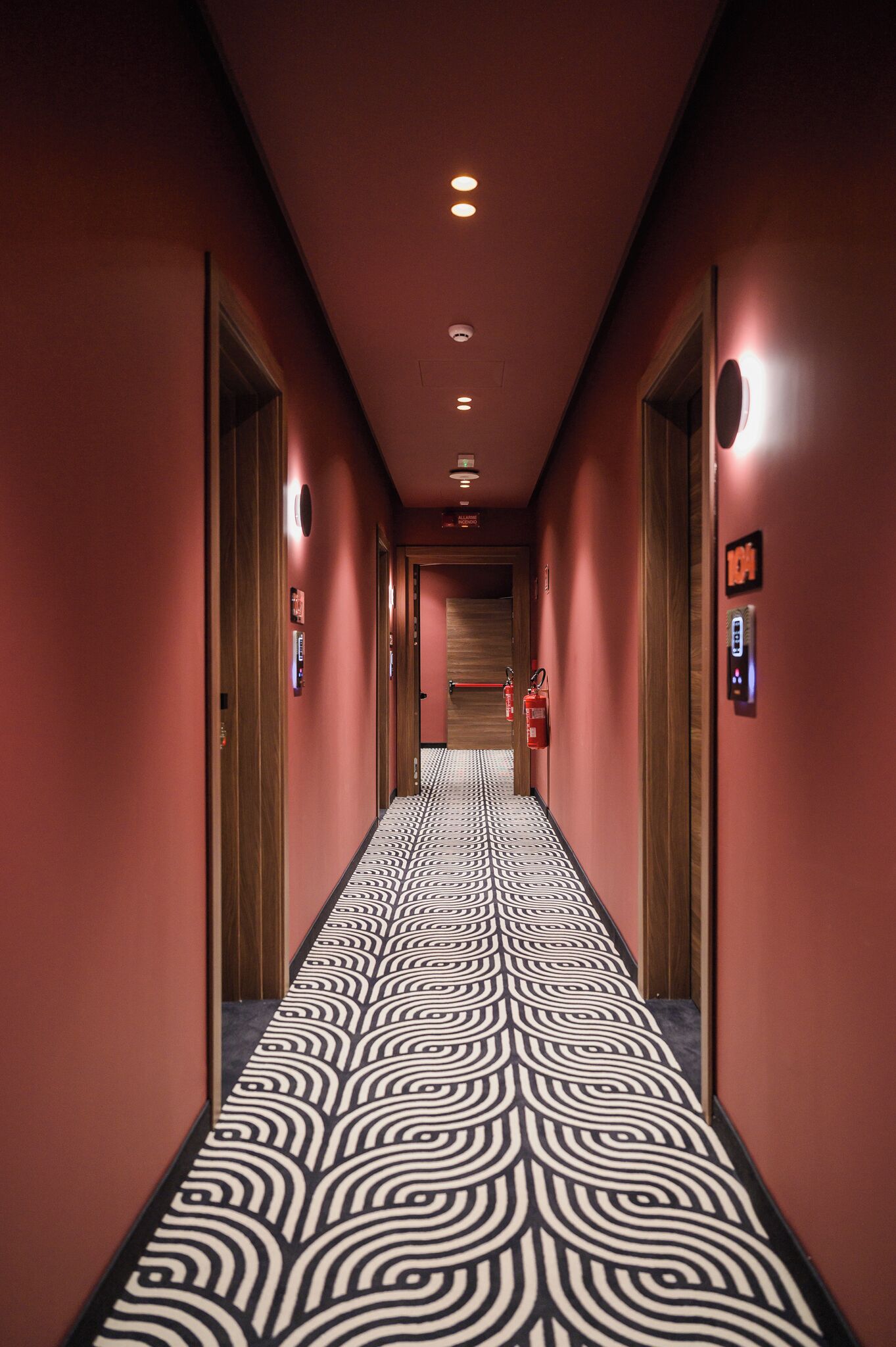Un pasillo de hotel con alfombra estampada, puertas de madera y un extintor.