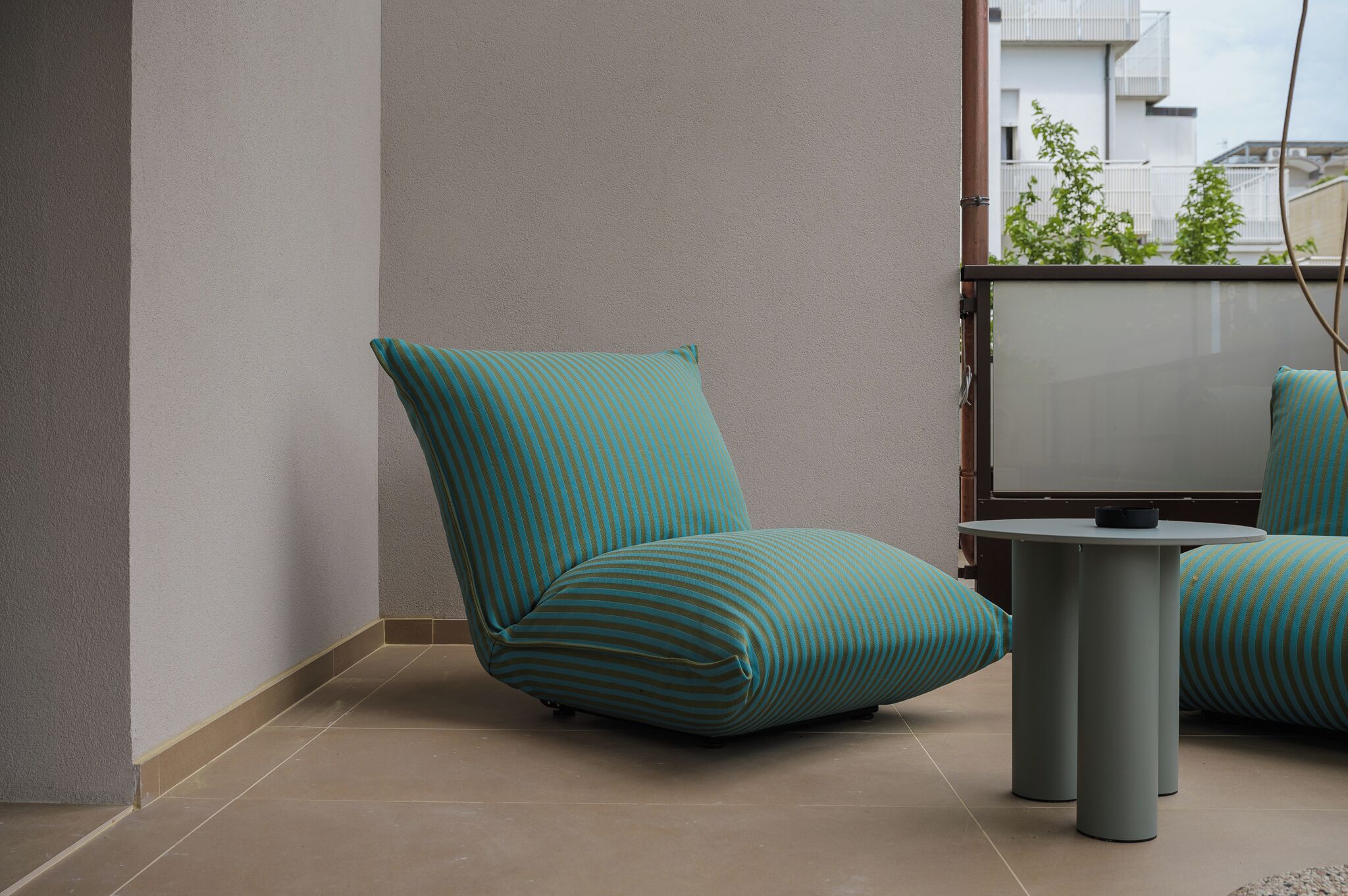 A corner of a room with a teal-striped chair, a small round table, and a view of a balcony with greenery.