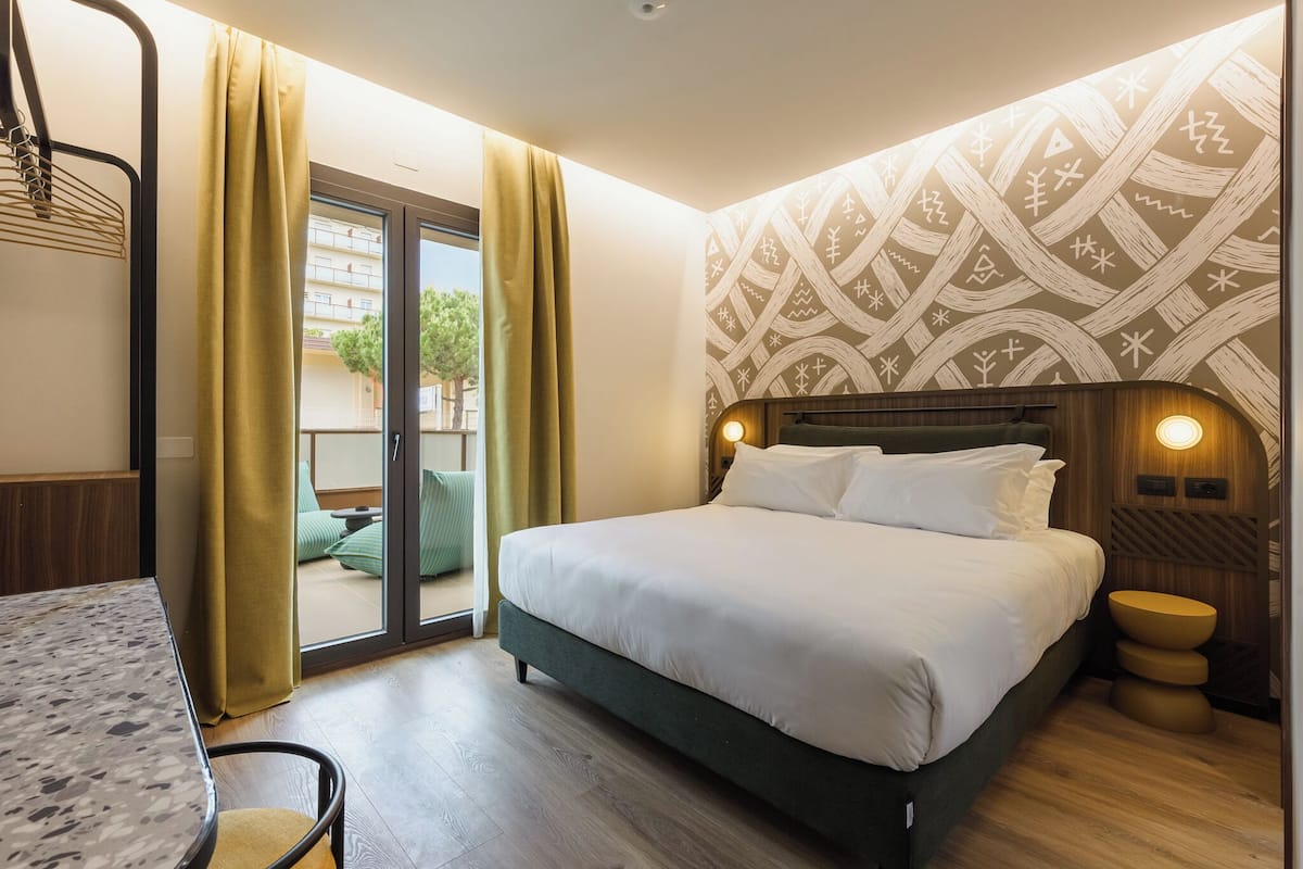 Une chambre d’hôtel avec un lit, un bureau, une chaise, une fenêtre avec des rideaux et un mur décoré de motifs.