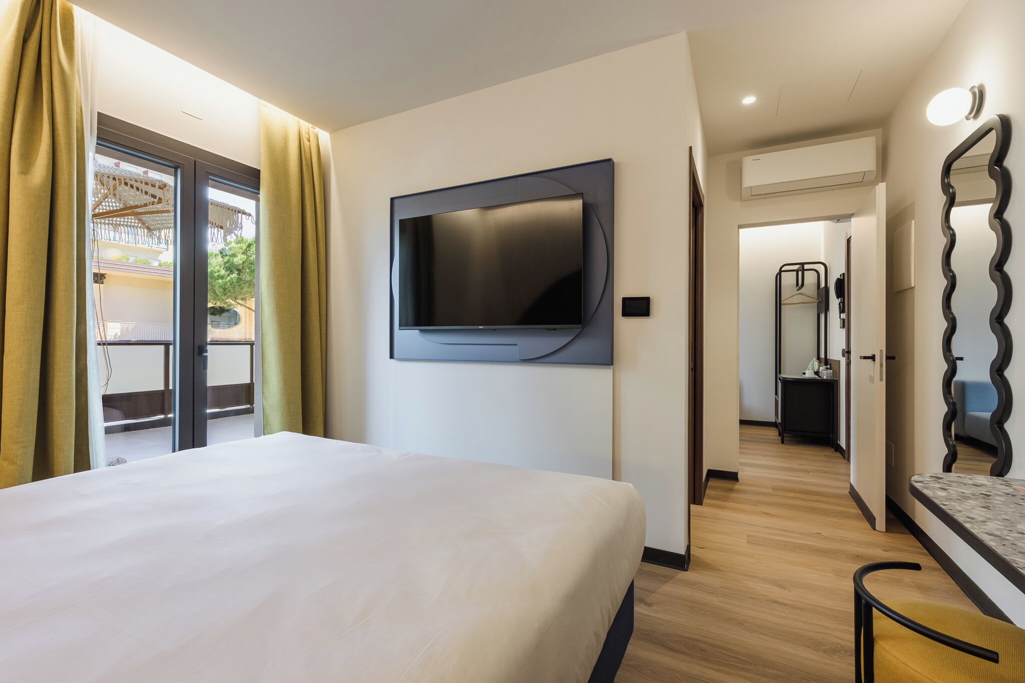 Habitación de hotel con cama, televisor montado, baño con espejo y vista al exterior.