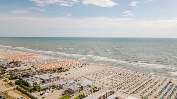 A beach with rows of umbrellas and sun loungers, a parking area, and buildings with signage.