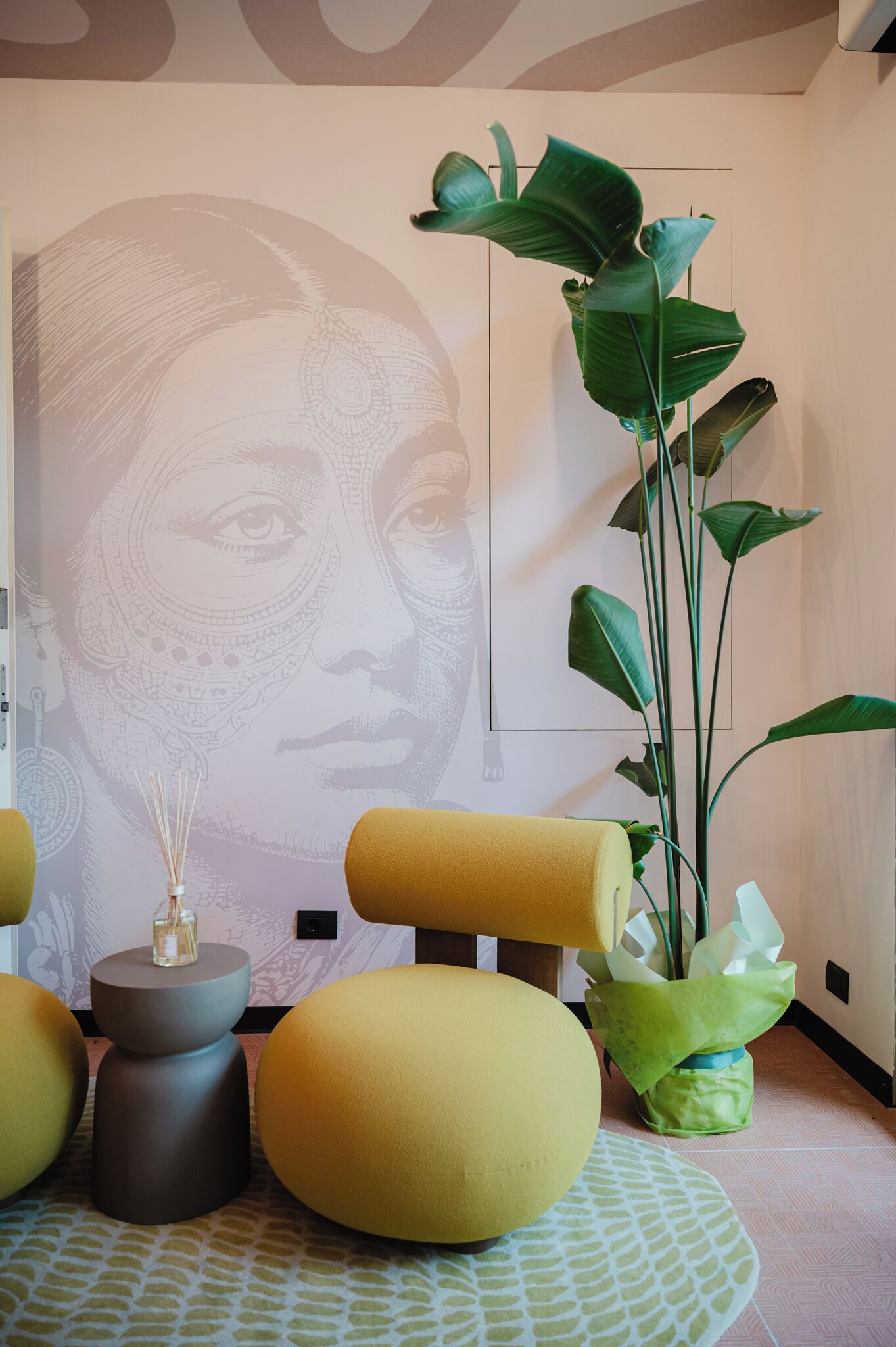 A room with a large wall mural, a tall green plant, yellow and grey furniture, and a patterned floor.