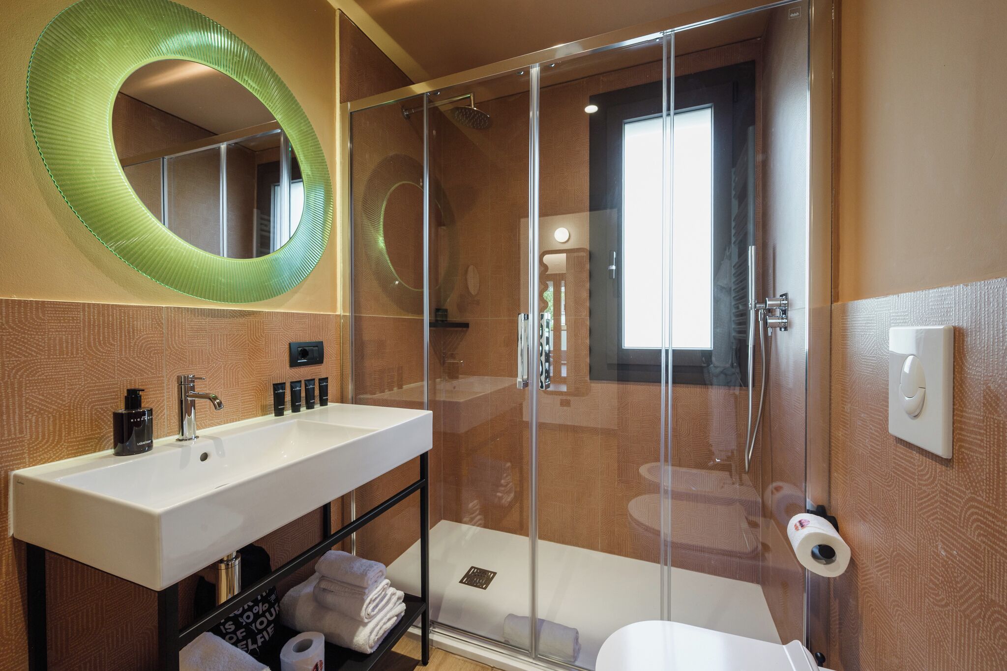 A bathroom with a glass-enclosed shower, a white sink, and a large mirror with a green circular frame.