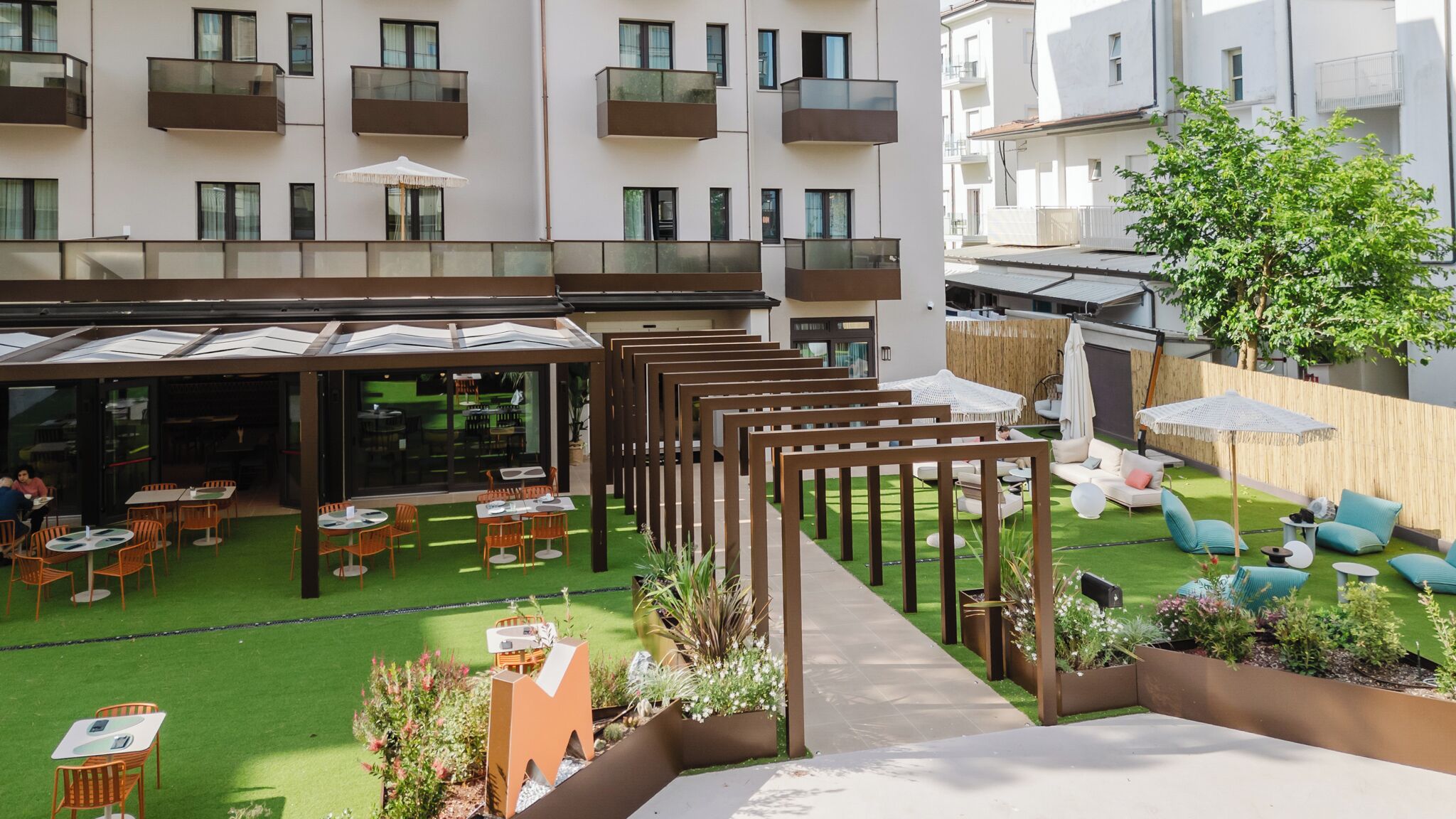 An outdoor patio with tables and chairs, a grassy area, and a building with balconies in the background.