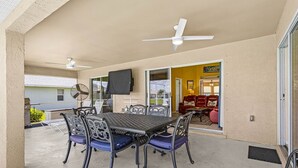 Un patio cubierto con una mesa, sillas, un ventilador de techo y un ventanal corredero que da a un espacio interior con un sofá y una televisión.