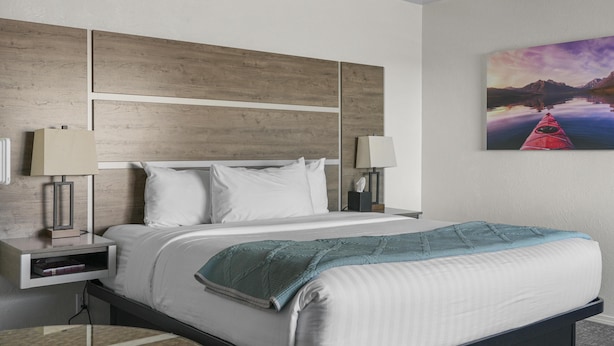 Ein modernes Hotelzimmer mit einem hölzernen Kopfteil, einem großen Bett mit weißer Bettwäsche, zwei Nachttischlampen und einem Gemälde von einer Berglandschaft.