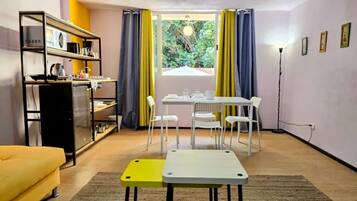 A compact living space with a kitchenette, a dining area with a table and chairs, a sofa, and a view through a window with curtains.