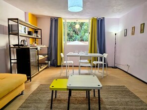 Un espace de vie compact comprenant un coin cuisine, un coin repas avec une table et des chaises, un canapé et une vue sur l’extérieur à travers une fenêtre avec des rideaux.