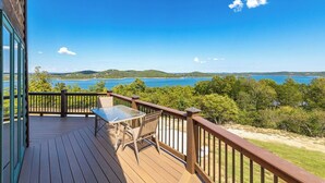 Eine Holzterrasse mit einem Glastisch und Stühlen, mit Blick auf einen See und Hügel.
