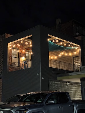 A two-story building with a garage, a balcony with a railing, and string lights.