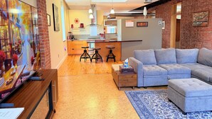 Una sala moderna con un sofá gris, un tapete azul estampado y una mesa de centro de madera. Hay una zona de cocina con encimera de madera y una lámpara colgante.