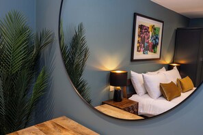 Un espejo curvo que refleja una cama con almohadas blancas y marrones, una mesita de noche con una lámpara y una obra de arte enmarcada en la pared.