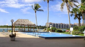 A pool area with a thatched-roof gazebo, palm trees, and a hotel building in the background. - Paradise on the Isles - Stunning Waterfront Views - Pool & Dock Steps Away (Naples)