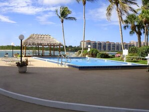 Ein Poolbereich mit einem Gazebo mit Reetdach, Palmen und einem Hotelgebäude im Hintergrund.