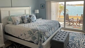 A bedroom with a bed, a nightstand, a lamp, a window with a view of the sea, and a balcony with chairs. - Paradise on the Isles - Stunning Waterfront Views - Pool & Dock Steps Away (Naples)