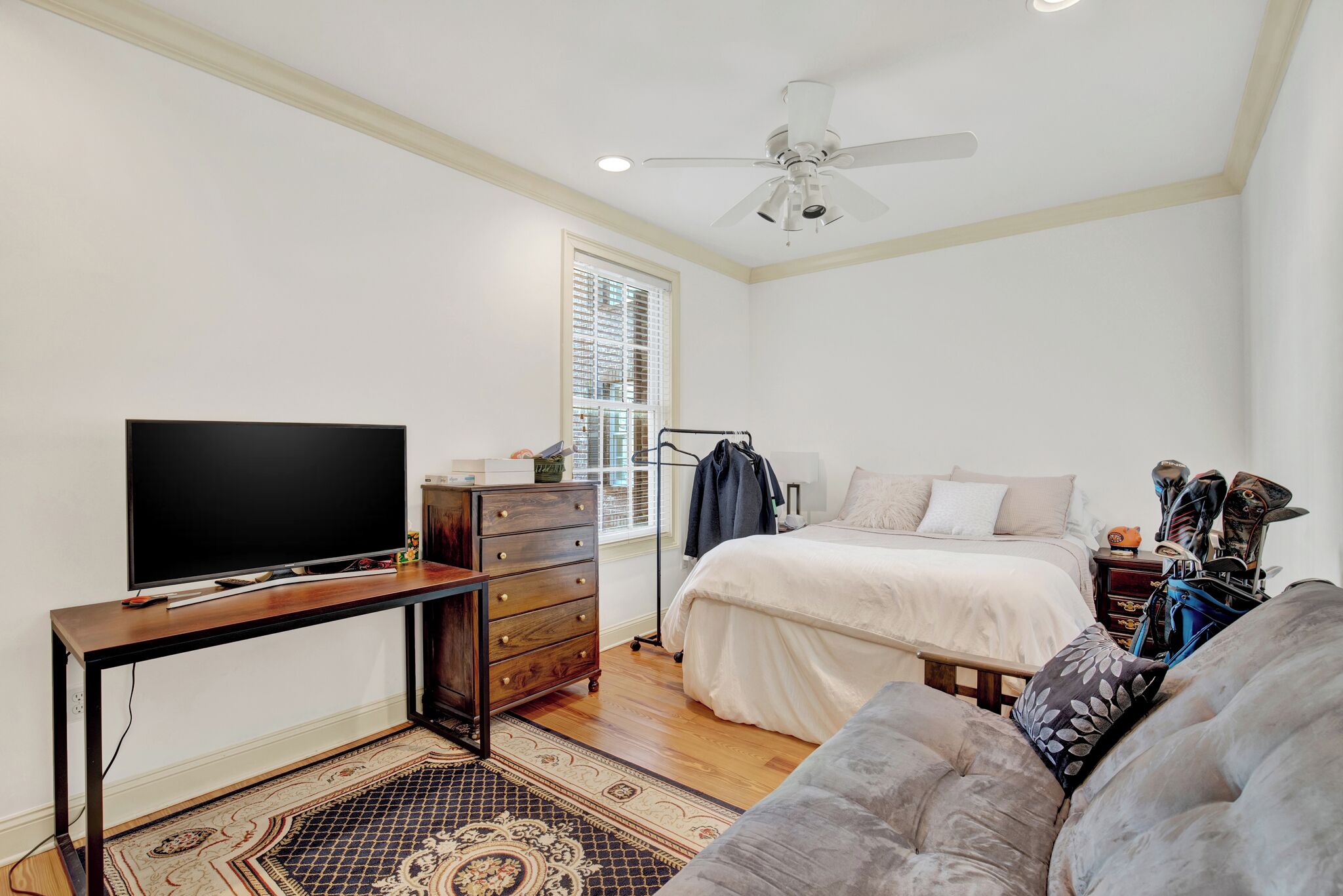 A bedroom with a bed, a TV, a dresser, a ceiling fan, and a window with blinds.