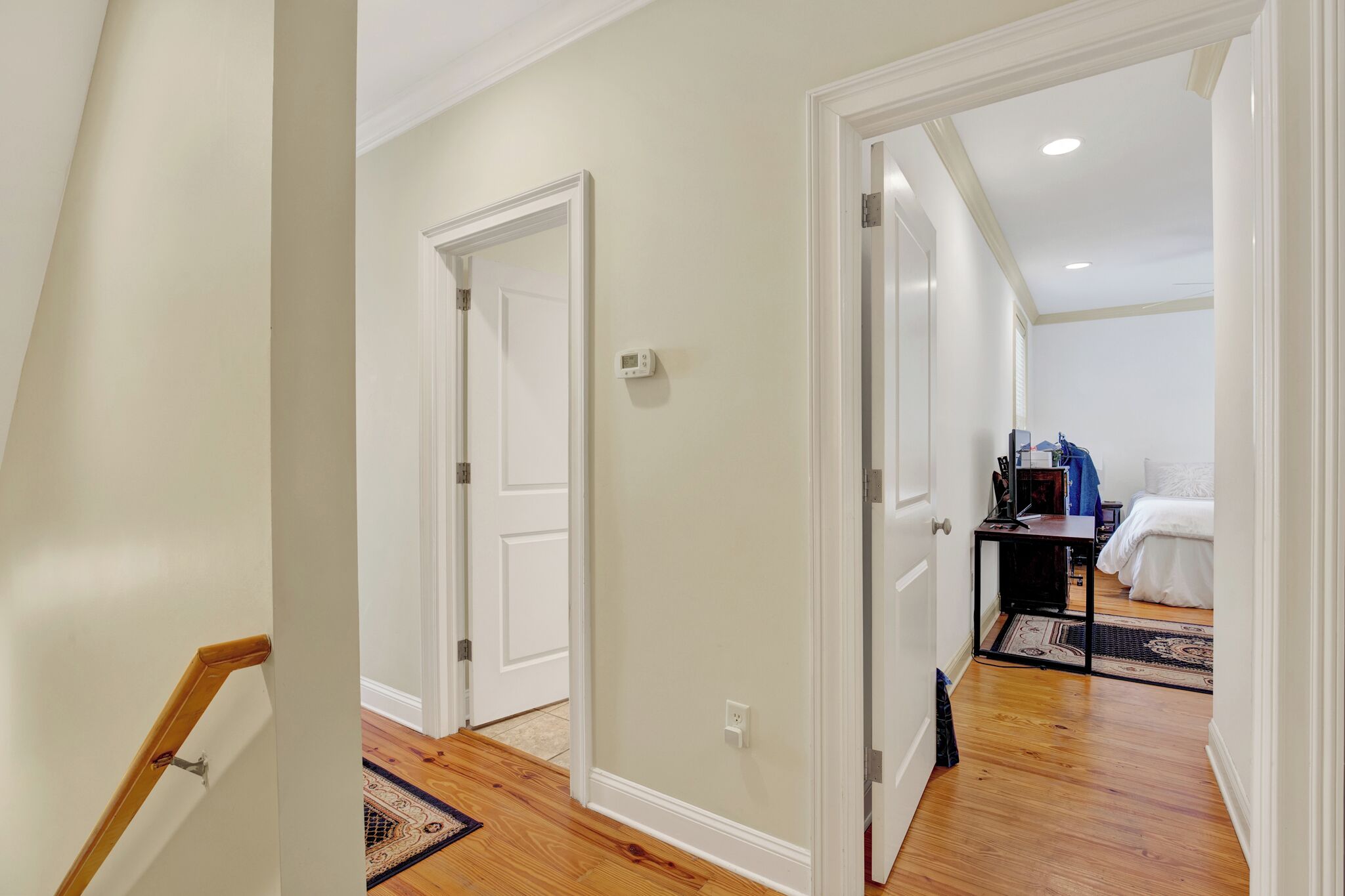 A hallway with wooden flooring, white walls, and a door leading to a bedroom with a bed and a nightstand.