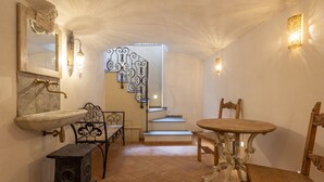 Una habitación con un lavamanos de piedra, una estufa pequeña, una mesa de madera y una escalera con barandillas de metal decorativas.